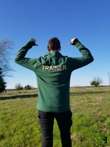 Person outdoors wearing a green jacket labeled TRAINER, showing thumbs up.