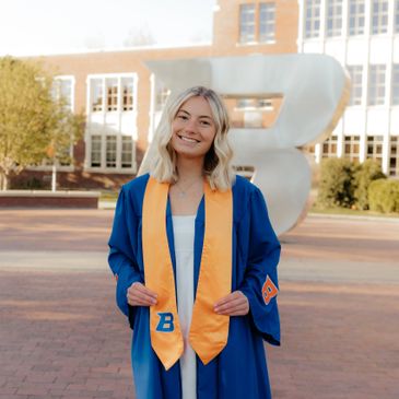 Graduate in blue gown and yellow stole standing outdoors with a large 'B' sculpture.