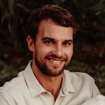 Smiling man with a beard wearing a light-colored shirt outdoors.