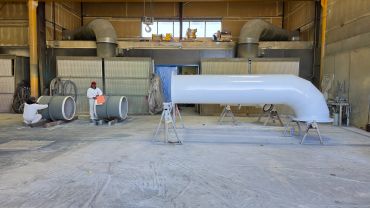 Workers painting large industrial pipes inside a spacious workshop.