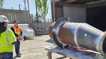 Workers examine a large industrial metal pipe outdoors.