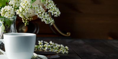 White flowers in a clear vase and a cup of tea in a saucer with a spoon on a table.