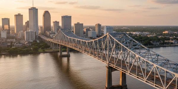 A steel bridge stretches over a river towards a city skyline at sunset.