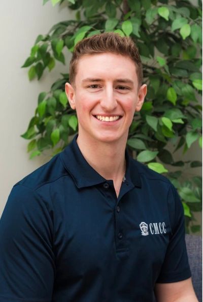 A young man in a navy blue polo shirt smiling in front of green plants.