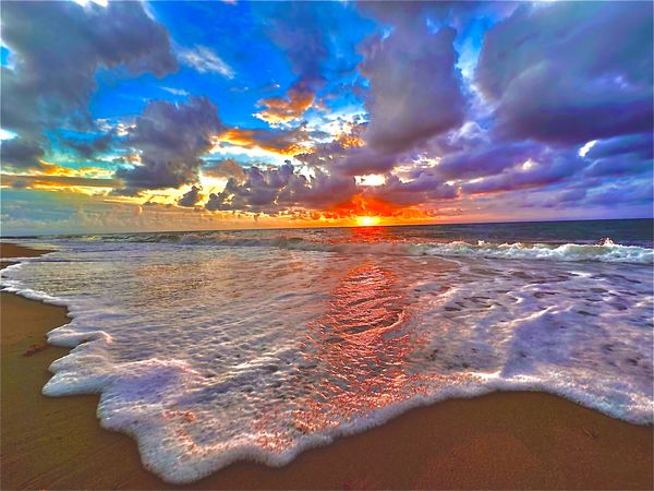 Sunset over ocean waves crashing on a sandy beach under a colorful sky.