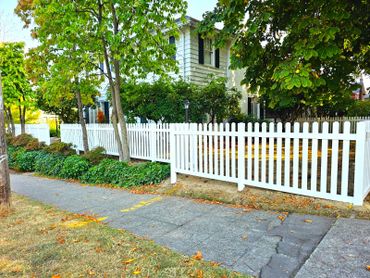 White picket fence around a suburban house with trees and sidewalk.