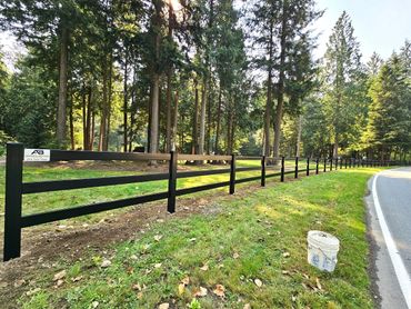 A black wooden fence runs alongside a road through a wooded area.