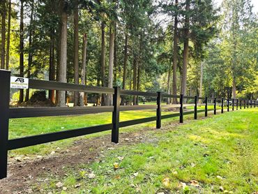 Black wooden fence lining a green grassy area with tall trees in the background.