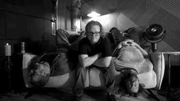 Three men in a dimly lit room, one sitting and two lying upside down on a couch.
