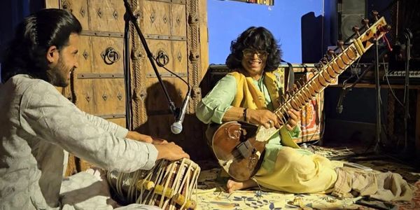 Two musicians playing traditional Indian instruments sit on the floor.
