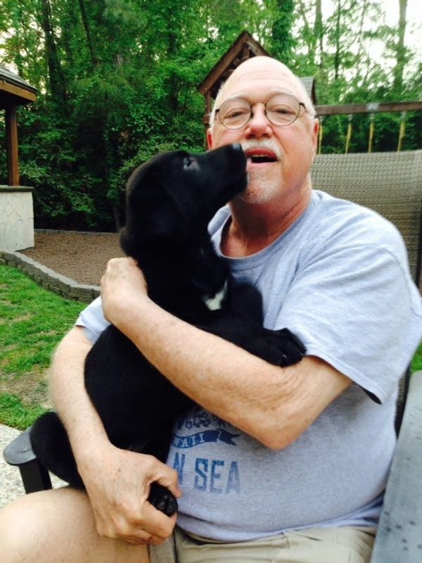 Man hugging a black puppy outdoors.