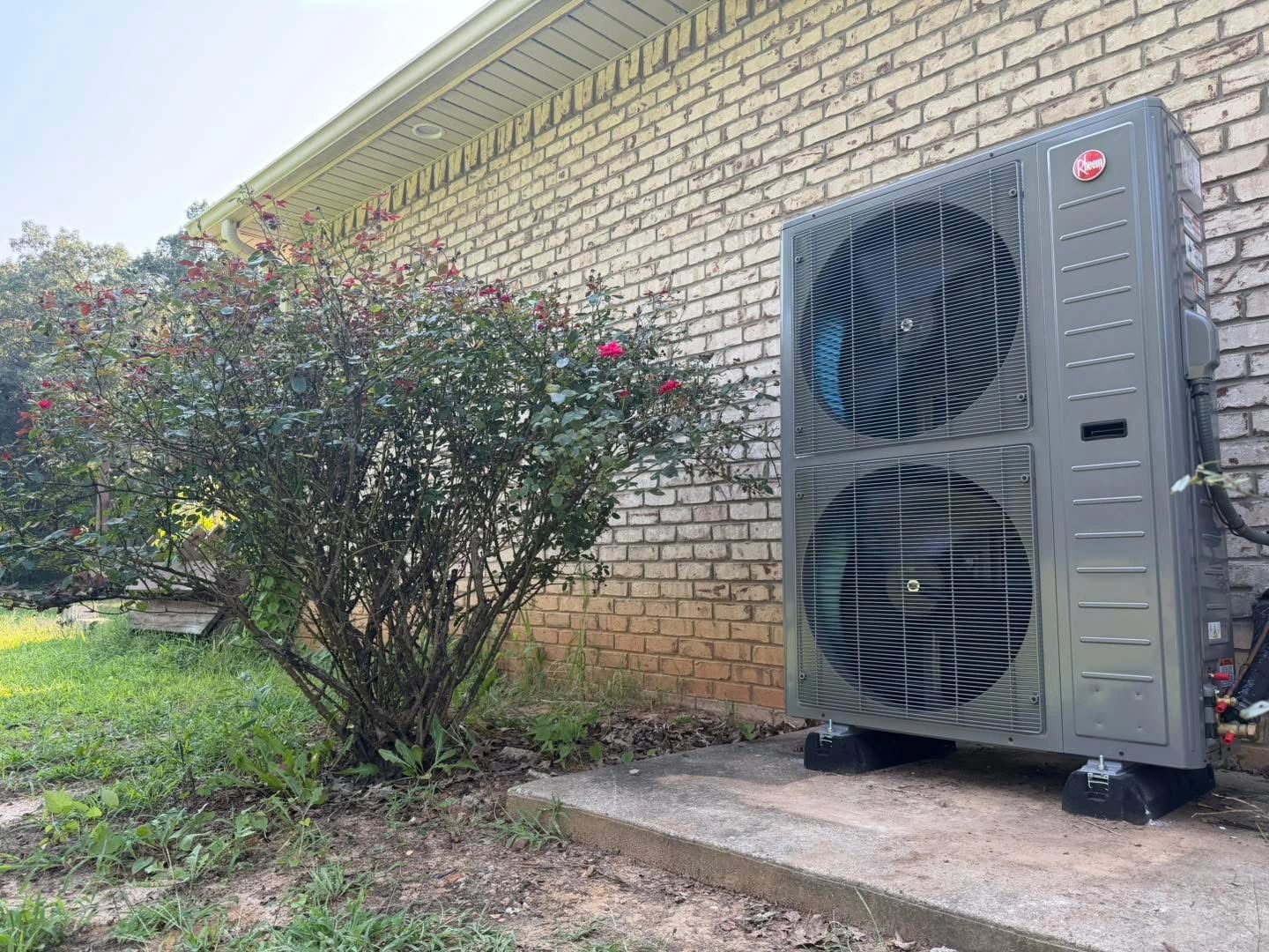 Outdoor air conditioning unit next to a blooming bush by a brick house.