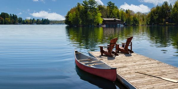 A peaceful lake scene with a dock, two chairs, and a canoe on a sunny day.