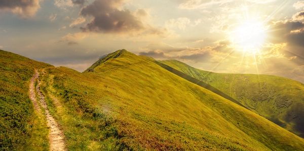 A sunlit mountain path stretches along green rolling hills under a partly cloudy sky.