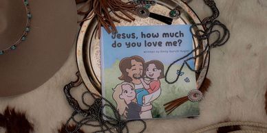 A children's book titled 'Jesus, how much do you love me?' surrounded by necklaces and a hat.