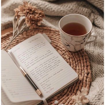 Cozy setup with a notebook, pen, dried flowers, and a cup of tea on a woven mat.