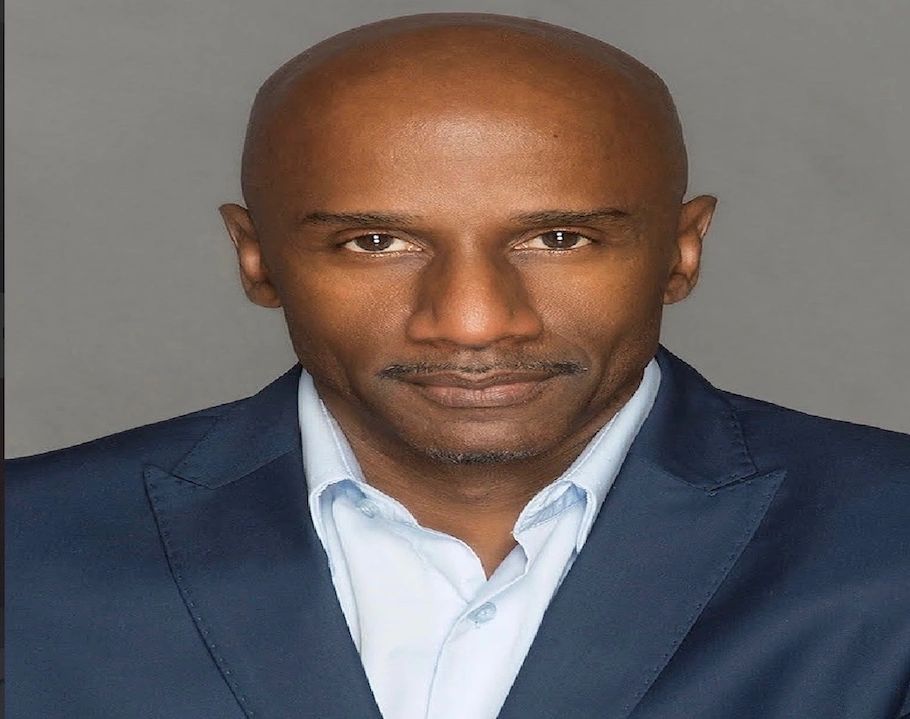 Confident man in a dark blue suit and light blue shirt against a gray background.
