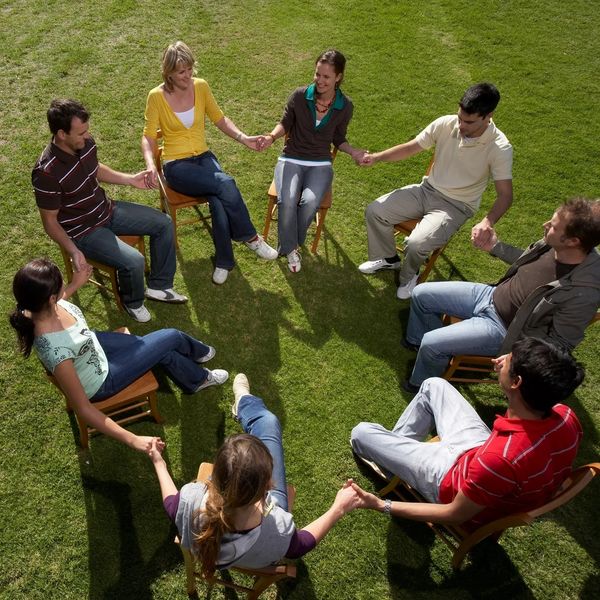 8 people sitting in a circle on grass holding hands, holding space for one other and others