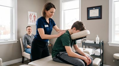 Massage Therapist providing pediatric massage to a seated youth client in a clinic with parent prese