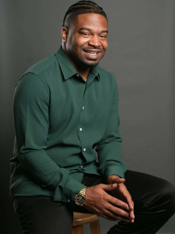 Smiling man in a green shirt sitting on a stool against a gray background.