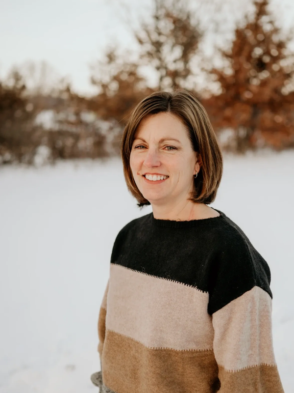 Woman smiling outdoors in a winter setting with snow and autumn trees.