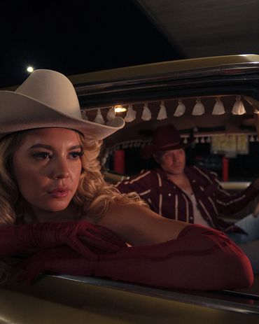 Woman in cowboy hat and gloves leaning out of car window at night.