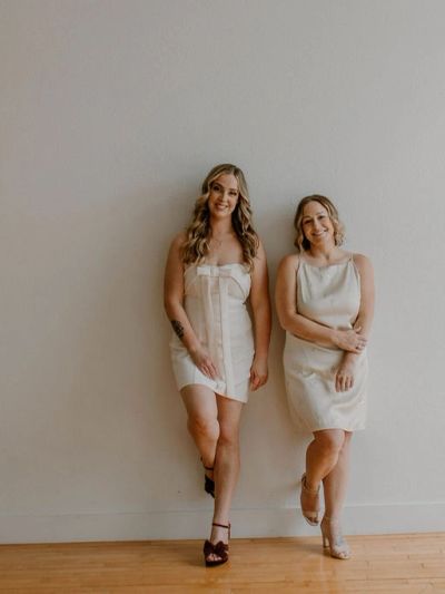 Two women in white dresses posing against a plain wall.