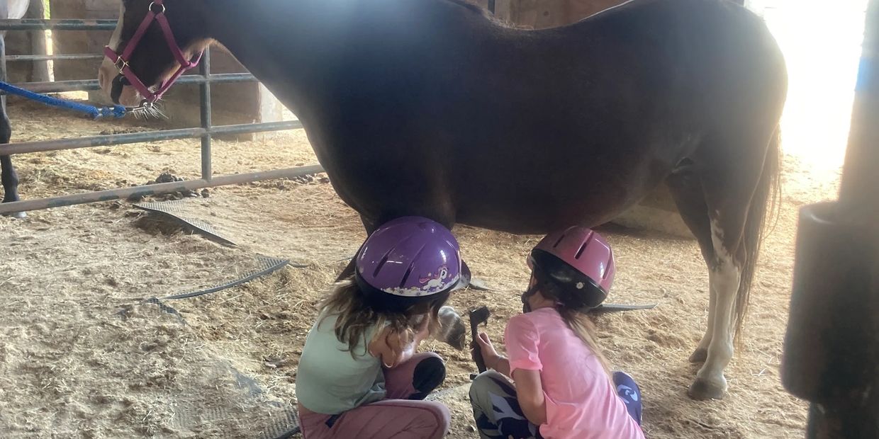 Two children with helmets near a horse in a stable.