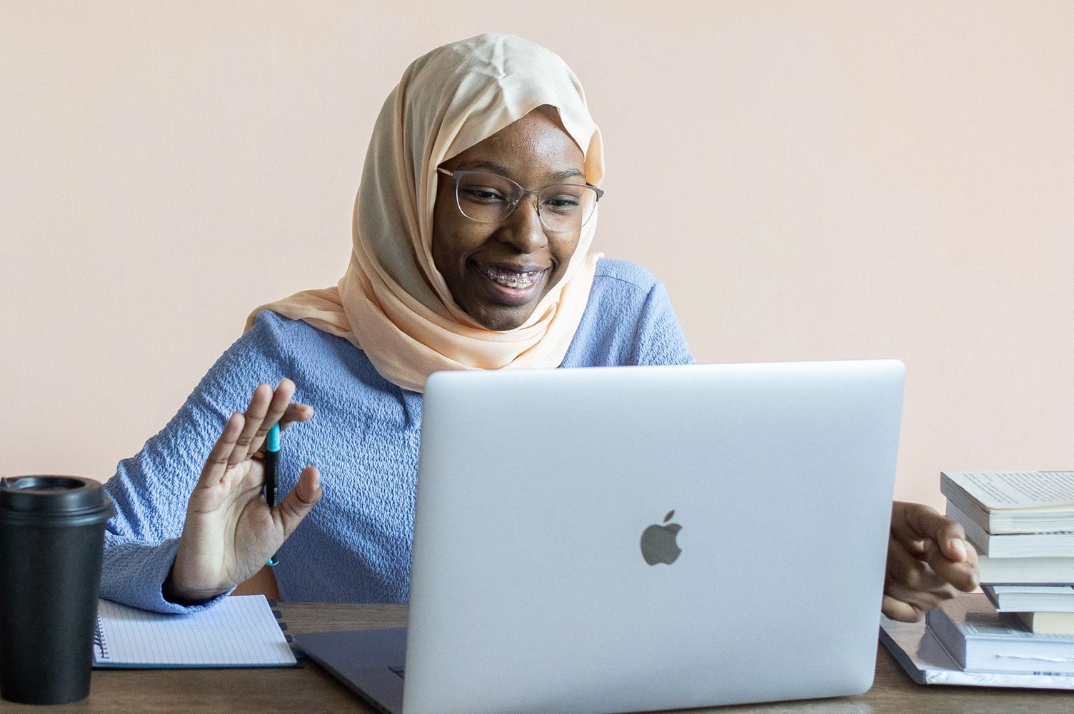 A smiling woman with a hijab sits in front of a laptop and waves at the screen.