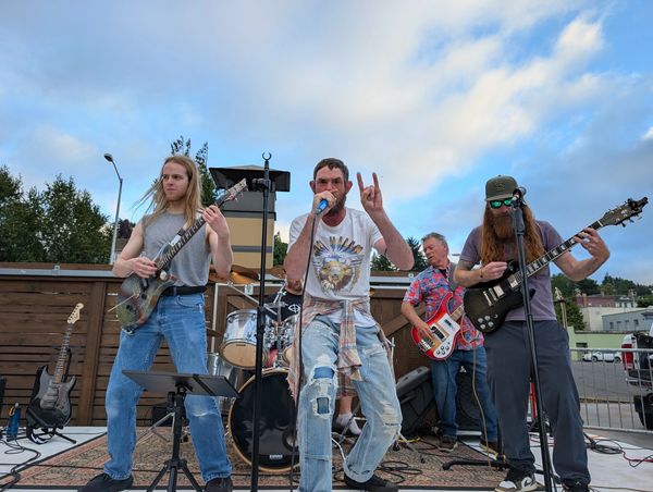 A band performing energetically outdoors with guitars and drums under a blue sky.