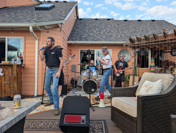 A band performing outdoors in a backyard setting with instruments and microphones.