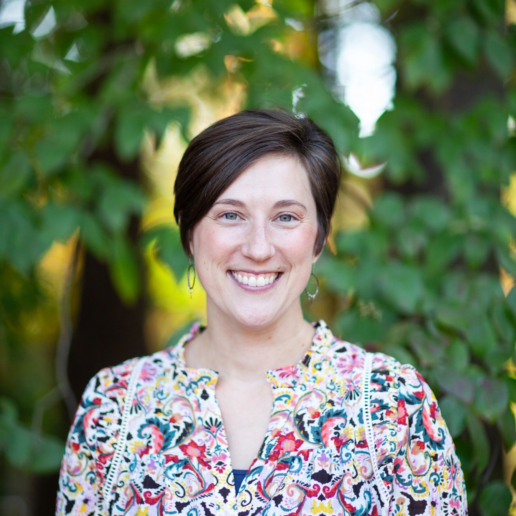 Smiling woman in a colorful patterned blouse stands outdoors with greenery in the background.