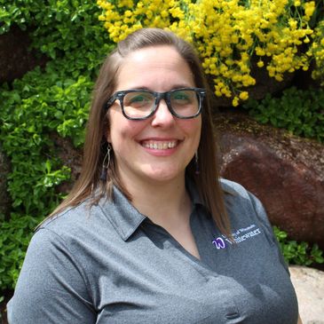 A head shot of a white woman with brown hair wearing a grey polo and rimmed glasses and smiling