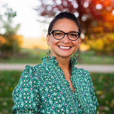 A head shot of a Brown woman wearing a green floral dress and black-rimmed glass and smiling