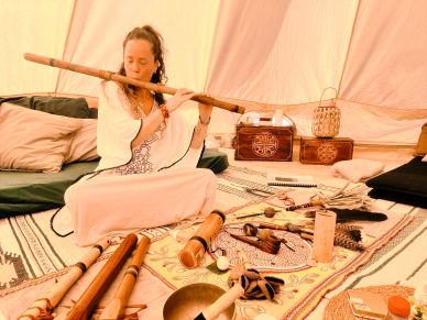 A woman playing a wooden flute inside a cozy tent with cushions and various objects around.