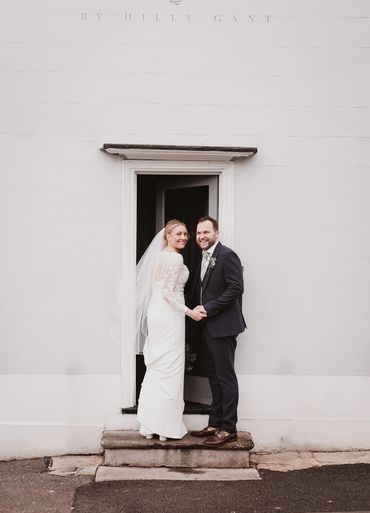 A newly wed couple looking at the camera holding hands at the door of The house by Hilly Gant 