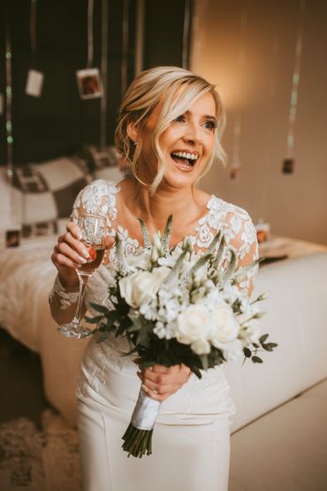 A stunning bride holding a Champlain flute and flowers smiling in her wedding dress at Friern Manor 
