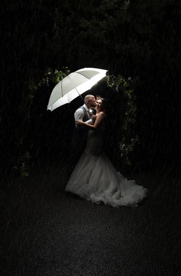 Groom kissing bride on cheek under an umbrella,  out the front of Stock Brook Manor, Essex. 
