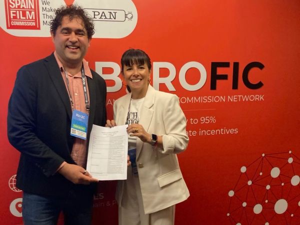 Dos personas sonrientes posan con un documento frente a un fondo rojo.