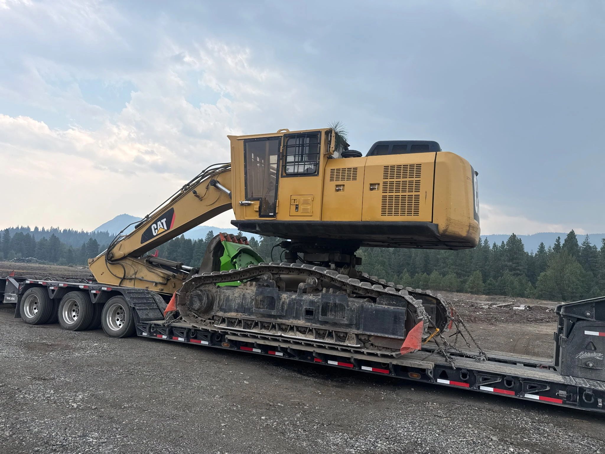 Yellow CAT excavator on a trailer with mountainous forest background.