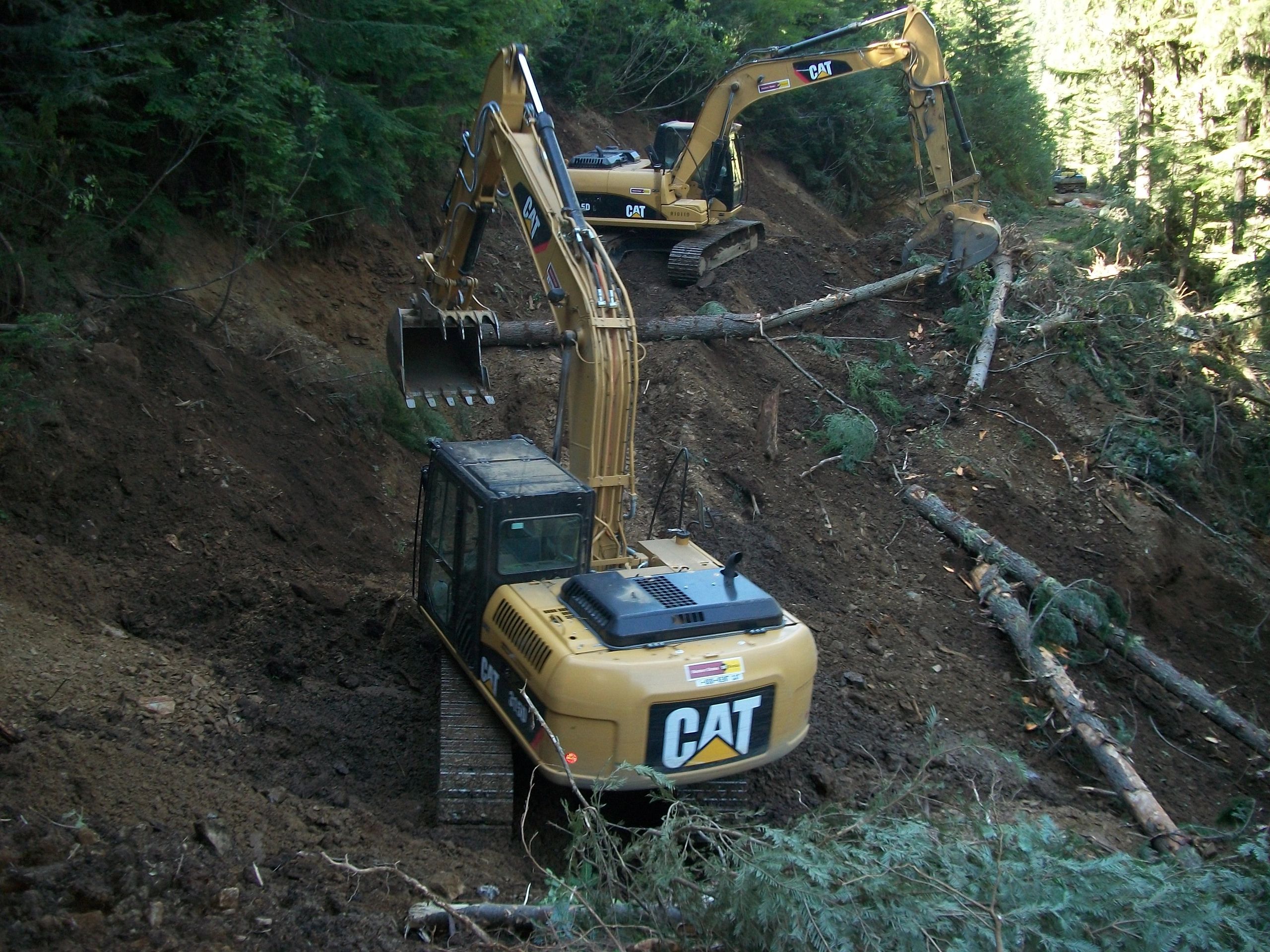 Two excavators clearing a forest path, moving logs and soil.