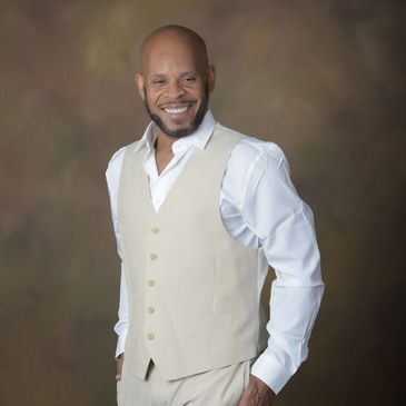 Smiling man in a beige vest and white shirt against a brown backdrop.