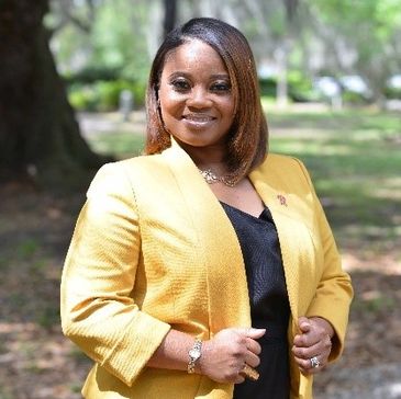 Confident woman in a yellow blazer smiling outdoors with greenery in the background.