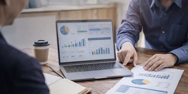 Two people analyzing business charts on a laptop and paper at a wooden table.