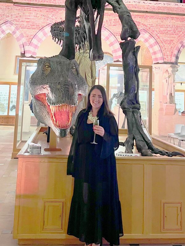 Woman in black dress posing with a dinosaur skeleton at a museum event.