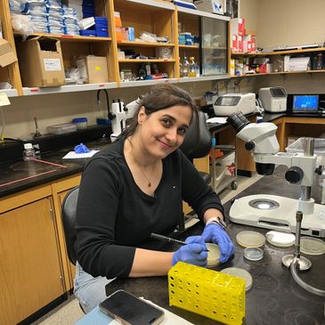 A woman in a lab works with petri dishes while smiling at the camera.