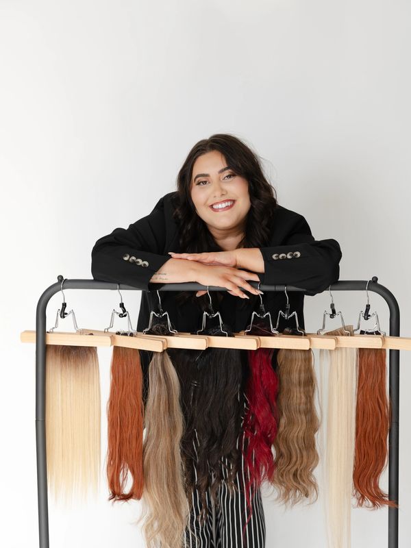 Woman smiling behind a rack of diverse hair extensions in various colors and textures.
