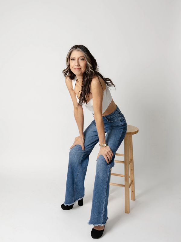 Woman in white crop top and jeans sitting on a wooden stool against a plain background.