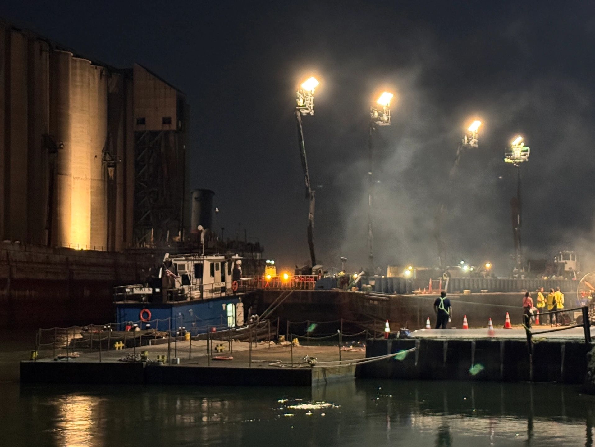 Nighttime industrial dock with workers and bright floodlights.