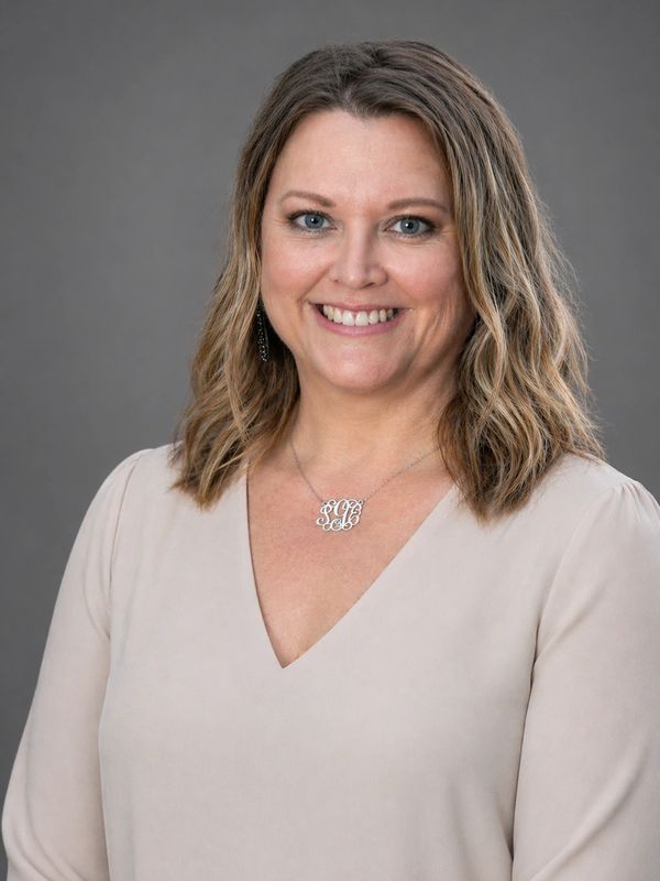 Smiling woman with wavy hair wearing a beige blouse against a gray background.